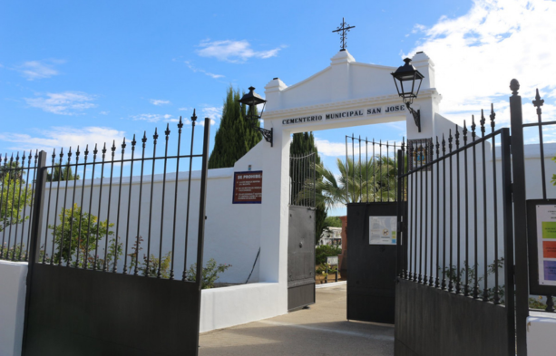 La foto captura la entrada al Cementerio Municipal San José ubicado en Villamanrique de la Condesa, Sevilla. Se presenta un portal de acceso blanco adornado con una cruz en la cima y dos estructuras en forma de campanario a los lados, lo que le confiere un aspecto tradicional y solemne. El portón es de metal, pintado de negro, con picos decorativos en la parte superior, y está parcialmente abierto, invitando a entrar. Justo al lado del portón, hay una placa informativa que indica la dirección del cementerio como la prolongación de la calle José Vélez, el número de teléfono para contacto y los horarios de verano e invierno, incluyendo horarios especiales para los meses de octubre y noviembre. Detrás de las puertas se observan palmeras y una variedad de vegetación, creando un contraste refrescante con la estructura blanca. El cielo azul claro con algunas nubes ofrece un fondo sereno. La foto transmite un ambiente de tranquilidad y respeto, elementos apropiados para un espacio destinado al recuerdo y la conmemoración. https://www.cementerio.info/