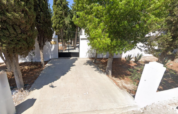 La foto muestra la entrada al Cementerio Municipal de Villanueva de San Juan en Sevilla. Está ubicada junto a la carretera A-406, en el kilómetro 31. En la entrada se ven dos pilares blancos que sostienen una puerta de metal con rejas blancas, que está cerrada. A cada lado de la entrada hay árboles que ofrecen sombra y verdor al entorno. El camino de acceso al cementerio está pavimentado con bloques de hormigón, y se puede observar un pequeño espacio de tierra a ambos lados del camino, con algunas plantas bajas y cactus. La presencia de árboles y vegetación sugiere un entorno tranquilo y natural. La luz del sol indica que la foto fue tomada durante el día y el cielo despejado sugiere un clima caluroso típico de la región. La imagen comunica una sensación de serenidad y es un ejemplo de la simplicidad y modestia que a menudo caracteriza a los cementerios en pequeñas localidades. https://www.cementerio.info/